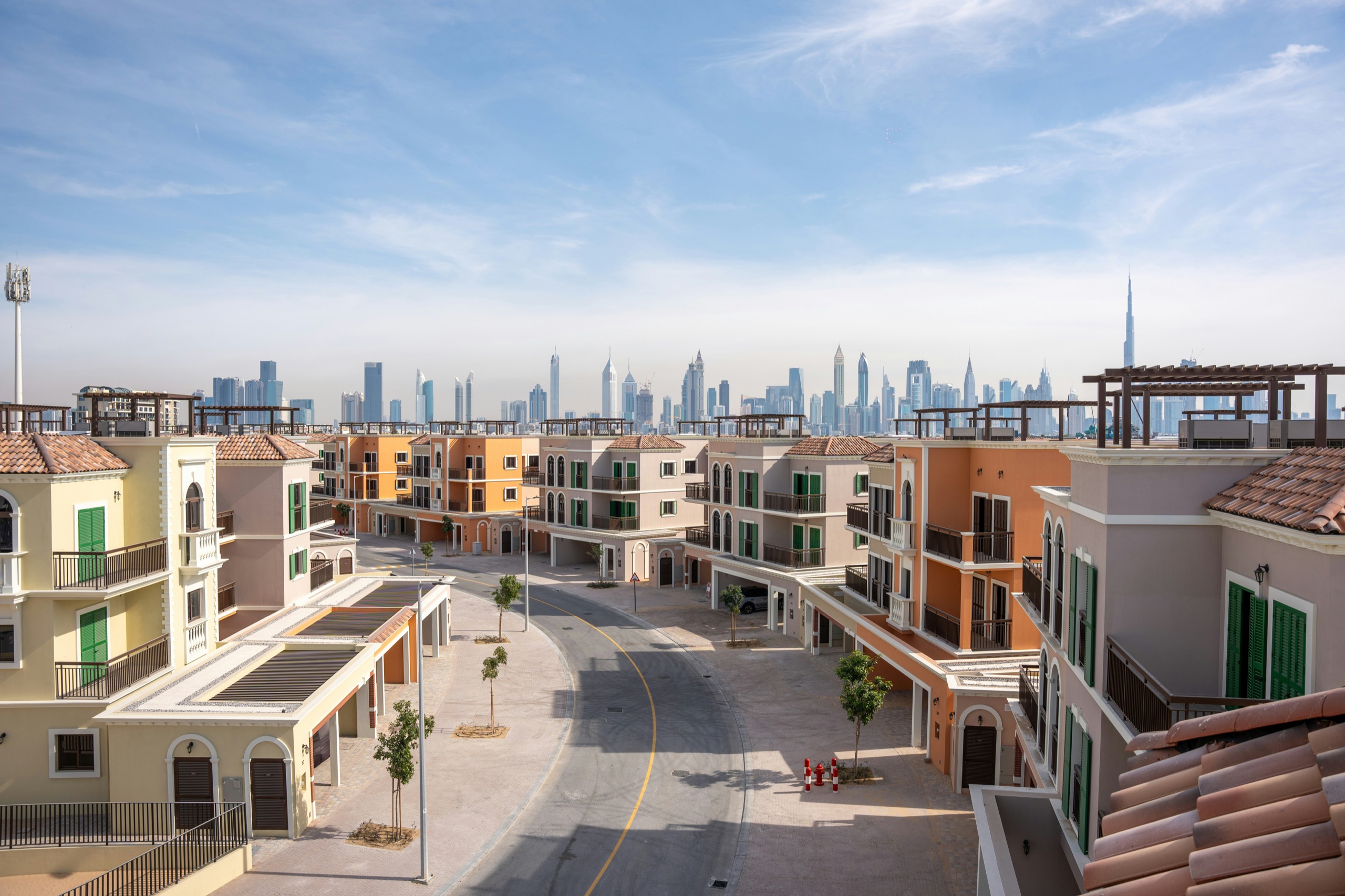 A street of pastel townhouses with terracotta roofs and roof terraces, the Dubai skyline behind